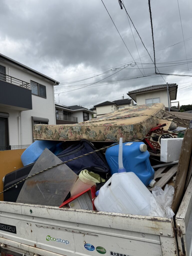 軽トラックの荷台にマットレス・ポリタンク・板材・スーツケースなどの不用品を積み上げ、ロープで固定した回収作業の様子