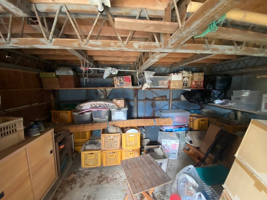 久山町の納屋で、棚や床に収穫用コンテナ、衣装ケース、木製家具、段ボールなどが保管されていた作業前の様子